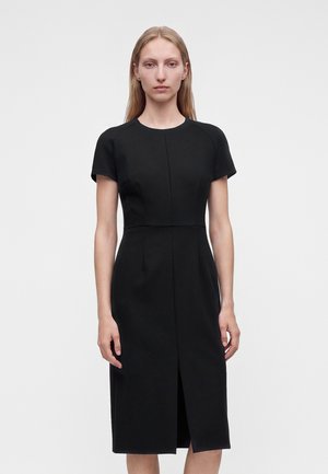 Femme aux longs cheveux blonds portant une robe noire cintrée à manches courtes avec une fente sur le devant, se tenant devant un fond clair uni.