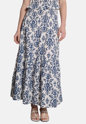 Biała długa spódnica maxi z niebieskim kwiatowym wzorem damaskowym, noszona przez osobę, w połączeniu z białymi sandałami na paseczki, przycięta na środku tułowia, widoczne stopy.