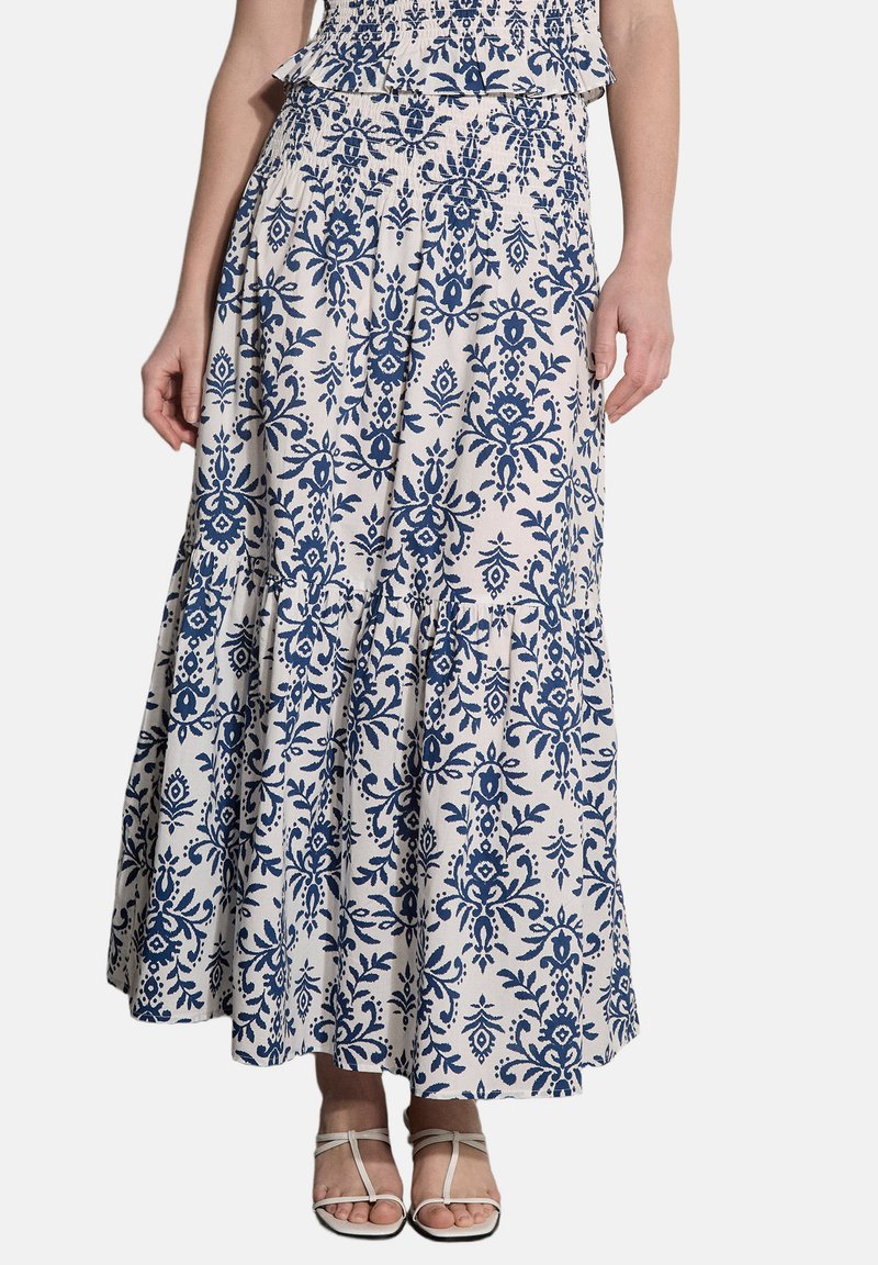 Jupe maxi blanche avec un motif floral damassé bleu portée par une personne, associée à des sandales blanches à lanières, cadrée au niveau du milieu du torse avec les pieds visibles.
