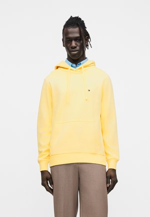 Mann mit geflochtenem Haar, der einen hellgelben Hoodie über einem blauen Hemd mit Hemdkragen und braunen Hosen vor einem schlichten weißen Hintergrund trägt.