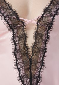 Pink satin garment with a deep V-neckline, accented by intricate black lace trim and frayed edges, featuring a crossed strap design.