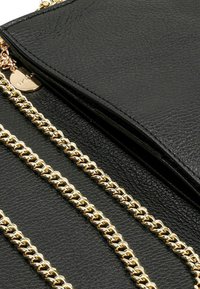 Borsa a tracolla nera in pelle con superficie testurizzata, dotata di una tracolla in catena dorata e un'etichetta metallica a forma di cuore. Chiusura con zip sulla parte superiore.