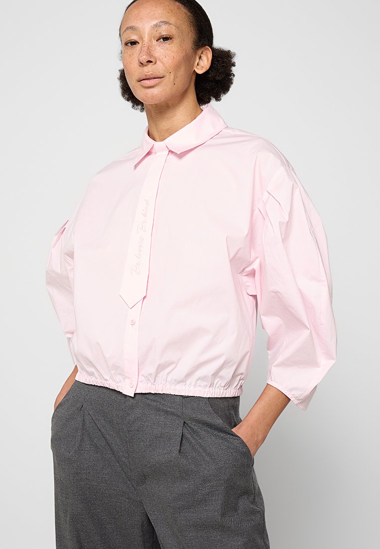 Camisa corta de color rosa claro con botones delanteros, cuello puntiagudo y dobladillo elástico. Cuenta con mangas anchas y abullonadas y detalles bordados sutiles.