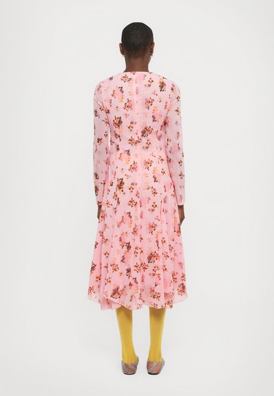 Langsleeve, jurk tot de knie met een roze bloemenpatroon op doorzichtige stof. Gecombineerd met gele panty’s en meerkleurige platte schoenen.