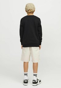 Felpa nera, pantaloni corti beige e sneakers neri con accenti bianchi. Vista da dietro, con una texture semplice e un cappellino.