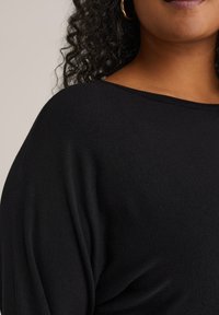 Fekete kötött pulóver széles nyakkivágással és laza szabással, puha textúrával és diszkrét redőzéssel a vállakon.