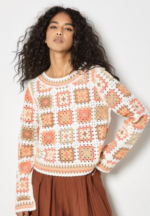 Žena s kudrnatými vlasy v bílém, broskvovém a hnědém háčkovaném svetru a rezavé plisované sukni, pózující s jednou rukou na boku.