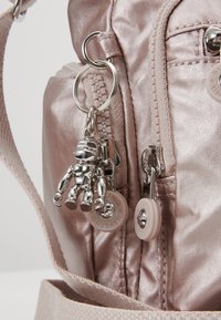 Metaliczny srebrny breloczek w kształcie figurki przypięty do jasnoróżowej błyszczącej torby, z widocznymi suwakami i okrągłymi, markowymi uchwytami suwaków.