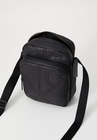 Black crossbody bag with quilted nylon material, featuring geometric stitching, a zip closure, and an adjustable strap. Compact and sleek design.