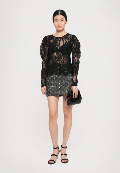 Femme portant un haut en dentelle noire transparente avec des manches bouffantes, une jupe mini en sequins argentés, tenant un sac à main noir texturé et des talons noirs à brides.