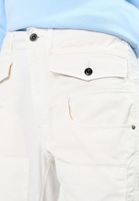 Pantaloni in cotone beige chiaro con due tasche frontali con chiusura a bottone e una texture liscia, abbinati a un top azzurro chiaro.