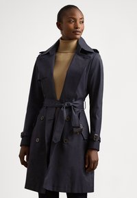 Μανώ στρατιωτικού τύπου σε ναυτικό μπλε, κατασκευασμένο από λείο ύφασμα, με ζώνη στη μέση, διπλό πέτο μπροστά και επιρράματα στους ώμους.