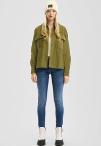 Camisa chaqueta de corduroy verde oliva con dos bolsillos frontales, combinada con una parte superior clara, jeans ajustados azul oscuro y un gorro de punto crema con logo.