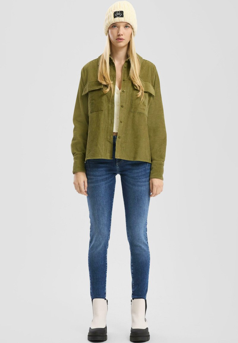 Camisa chaqueta de corduroy verde oliva con dos bolsillos frontales, combinada con una parte superior clara, jeans ajustados azul oscuro y un gorro de punto crema con logo.
