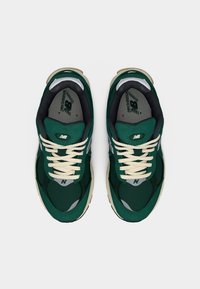 Ζευγάρι σκούρα πράσινα αθλητικά παπούτσια New Balance με μπεζ κορδόνια και μαύρη εσωτερική επένδυση, σε θέα από πάνω σε λευκό φόντο.