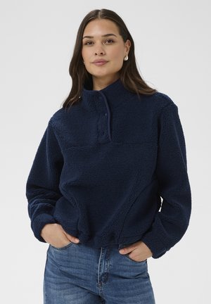 Mujer con un forro polar azul marino de cuello alto y botones de presión, con las manos en los bolsillos, sobre un fondo liso y claro.