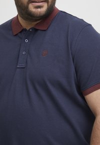 Marineblå polo skjorte med en maroon krage, som har tre knapper og et lite brodert logo på brystet. Myk, teksturert stoff.