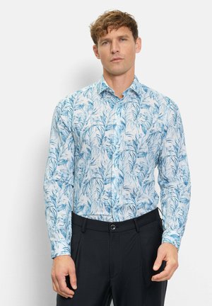 Mann trägt ein weißes Hemd mit blauen Blattmustern und schwarze Hose, steht vor einem einfarbigen hellen Hintergrund.