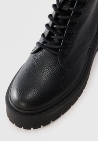 Bota de cuero negro con acabado texturizado, suela gruesa y cordones negros sobre un fondo blanco.