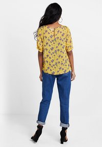 Blouse florale jaune à manches courtes bouffantes, associée à un jean en denim bleu retroussé. Des escarpins noirs accentuent la tenue.