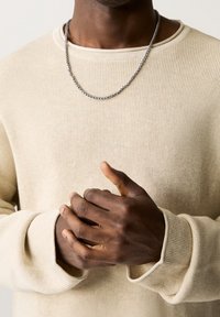 Collana di catena argentata poggiata su un maglione beige chiaro a coste. Le mani sono intrecciate, mostrando una pelle scura e unghie curate.