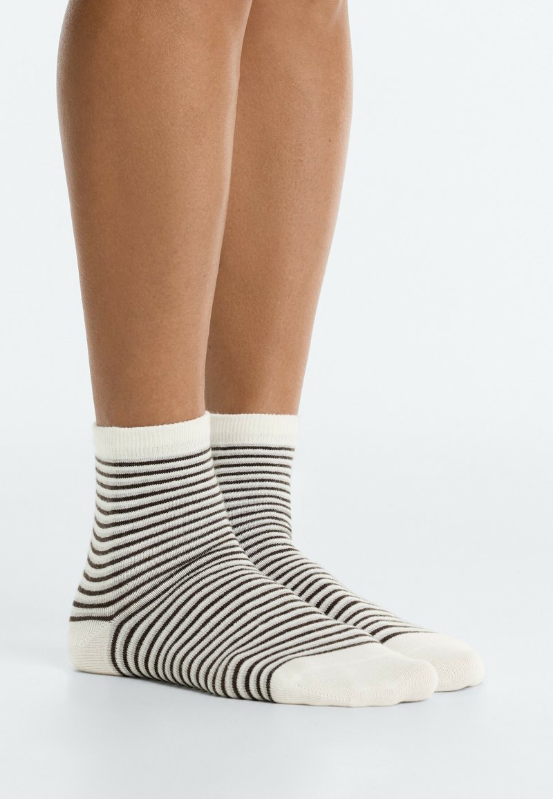Jambes portant des chaussettes mi-cheville à fines rayures horizontales noires et blanches, debout sur une surface blanche sur un fond uni.