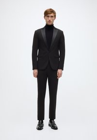 Zwart smokingjack met satijnen revers, gecombineerd met een zwarte coltrui. Slim-fit ontwerp en glanzende zwarte loafers. Effen achtergrond.