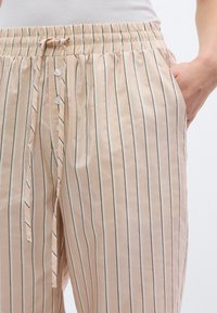 Ανοιχτό μπεζ παντελόνι με ρίγες, ελαστική μέση, κούμπωμα με κουμπί μπροστά και πλαϊνές τσέπες. Το ύφασμα έχει λεία υφή και διακριτική λάμψη.