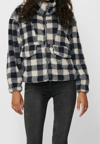 Veste en polaire avec col montant, présentant un motif à carreaux bleu marine et crème, fermeture éclair et deux poches devant. Tissu doux et texturé.