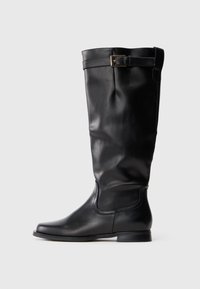 Bota de couro preta até o joelho com bico arredondado, salto baixo e uma faixa decorativa com fivela dourada perto do topo.
