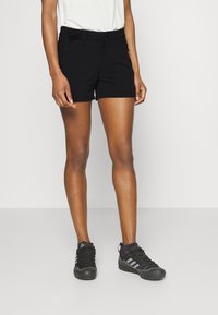 Person med svarta shorts, svarta Adidas-sneakers och en vit topp, står mot en enkel ljus bakgrund.