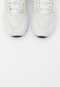 Nike sneakers with a light neutral upper, mesh texture, green accents, white laces, and a black and white sole.