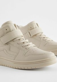 Baskets montantes beiges en cuir lisse avec une sangle texturée, un système de laçage et un bout perforé pour la ventilation.