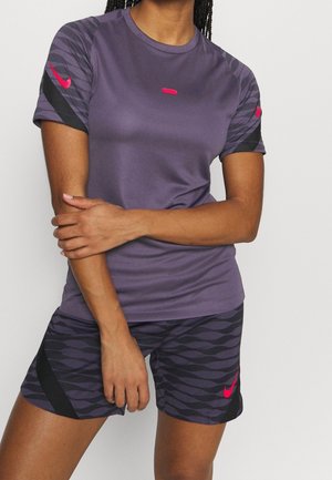 Vrouw draagt een paarse sport-t-shirt en gebloemde shorts met roze Nike-logo's, armen gekruist, tegen een effen achtergrond.