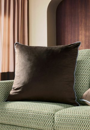 Großes dunkelbraunes Samtkissen auf einem grün-beigen, strukturierten Sofa mit beigefarbenem Vorhang und Holzwand im Hintergrund.