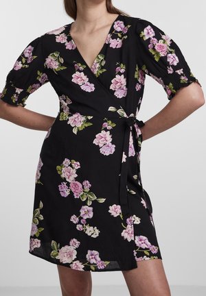 Czarna sukienka kopertowa z różowym i fioletowym kwiatowym nadrukiem, krótkimi bufkami na rękawach oraz wiązaniem z boku w talii, noszona przez osobę stojącą z rękami na biodrach.