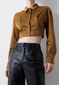 Donna che indossa una camicia corta marrone con bottoni e pantaloni neri in pelle trapuntata a vita alta, in piedi contro un muro bianco.