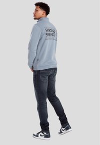 Ljusblå fleece-pullover med svart tryck på baksidan, kombinerad med mörka jeans och svart-vita sneakers.