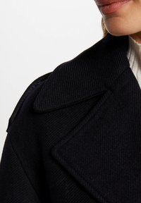 Marineblauw wollen jas met een gestructureerde afwerking, voorzien van een brede geknipte kraag en een getailleerde pasvorm. Close-up van de kraag en schouderdetails.