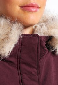 Burgundisk vinterjacka med en tjock fuskpälsluva, som har en mjuk tygstruktur och en hög krage för värme och skydd.