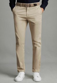 Chinos beige en coton lisse, dotés de poches latérales et d'une ceinture tressée. Associés à des baskets blanches, sur fond gris.