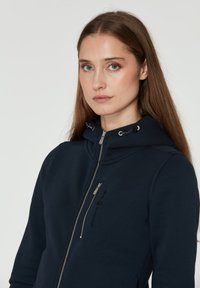 Sudadera con cremallera en azul marino, fabricada en tejido suave y con un cuello alto. Presenta una cremallera metálica y detalles de cordón en la capucha. Incluye logotipo en la parte frontal.