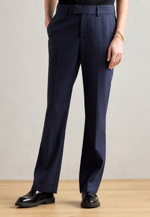 Person iført navyblå bukser med striber og lige ben, med sorte loafers og en sort top, står på et trægulv.