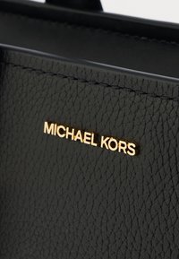 Schwarze, strukturierte Lederhandtasche mit einem goldenen "MICHAEL KORS"-Logo-Schild. Sie verfügt über eine elegante Nahtführung und ein modernes, klares Design, das Haltbarkeit und Stil betont.