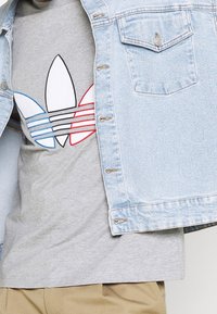 Ljusblå jeansjacka över en grå t-shirt med en flerfärgad Adidas-logotyp med svarta, röda och blå ränder, slät textur.