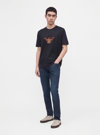 Млад мъж, облечен в черна тениска Emporio Armani, сини дънки и бежови маратонки, стоящ с една ръка в джоба си пред едноцветен фон.