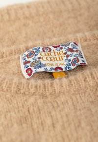 Beige wollen trui met geribde halslijn; heeft een kleurrijk label van stof met bloemenpatronen en de tekst "cache coeur". Maatlabel zichtbaar.