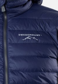 Dunkelblauer isolierter Pufferjacke mit horizontalen Steppmustern, silbernem Reißverschluss und "SWEDEMOUNT"-Logo über einer Berggrafik.