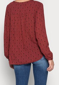 Frau trägt eine rostrote Bluse mit langen Ärmeln und schwarzem Fächer-Muster, kombiniert mit blauen Jeans, von hinten vor einem einfarbigen Hintergrund gezeigt.
