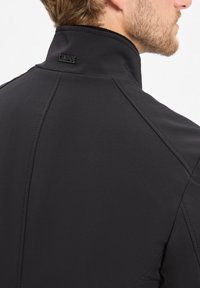 Zwarte softshelljas met een hoge kraag. Heeft een gestructureerd oppervlak en een verhoogd logo op de kraag, met zichtbare stikseldetails.